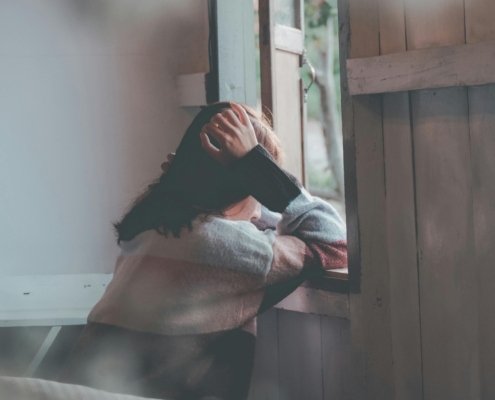 A person sitting indoors, leaning on a windowsill with one hand resting on their head, looking outside. The scene includes soft natural light, wooden panels, and hanging pinecones near the open window.
