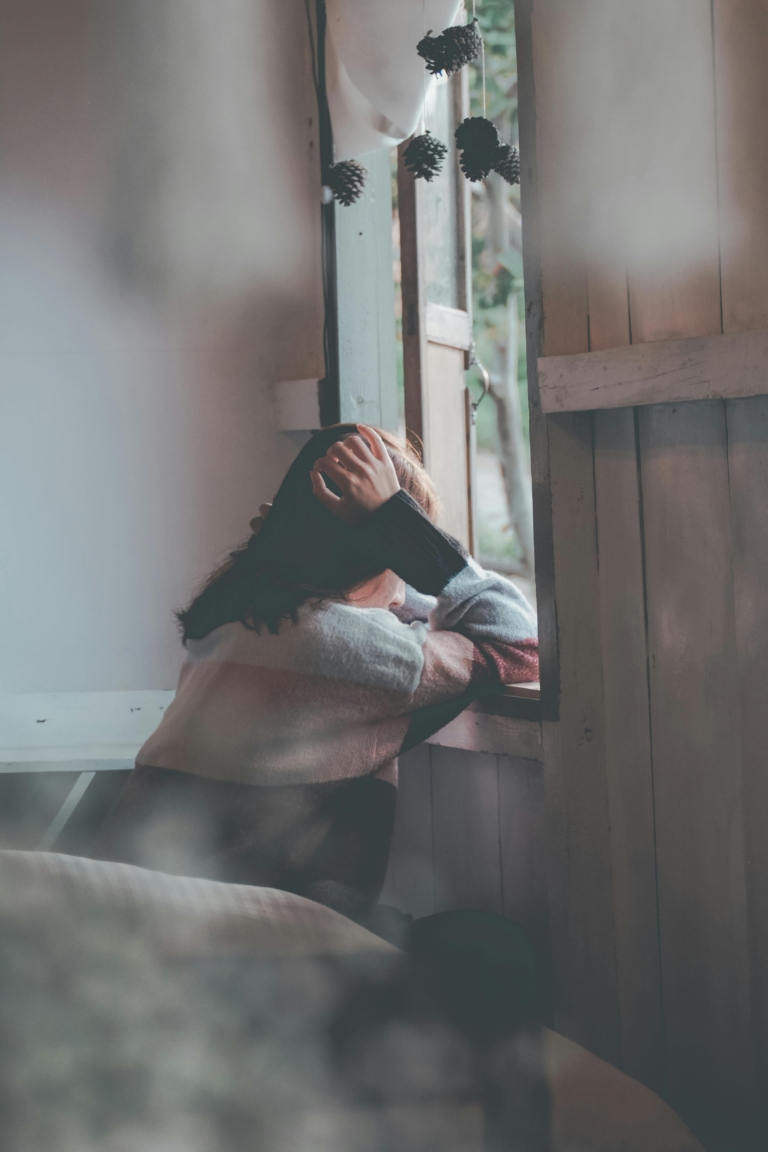 A person sitting indoors, leaning on a windowsill with one hand resting on their head, looking outside. The scene includes soft natural light, wooden panels, and hanging pinecones near the open window.