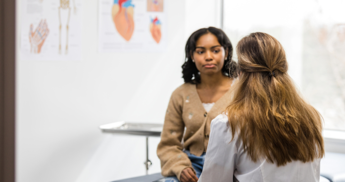An image of a women sitting on an examination bed listening to a female doctor sitting opposite her